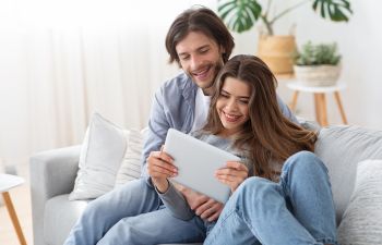 Happy man and woman sitting on couch at living room, holding digital tablet.