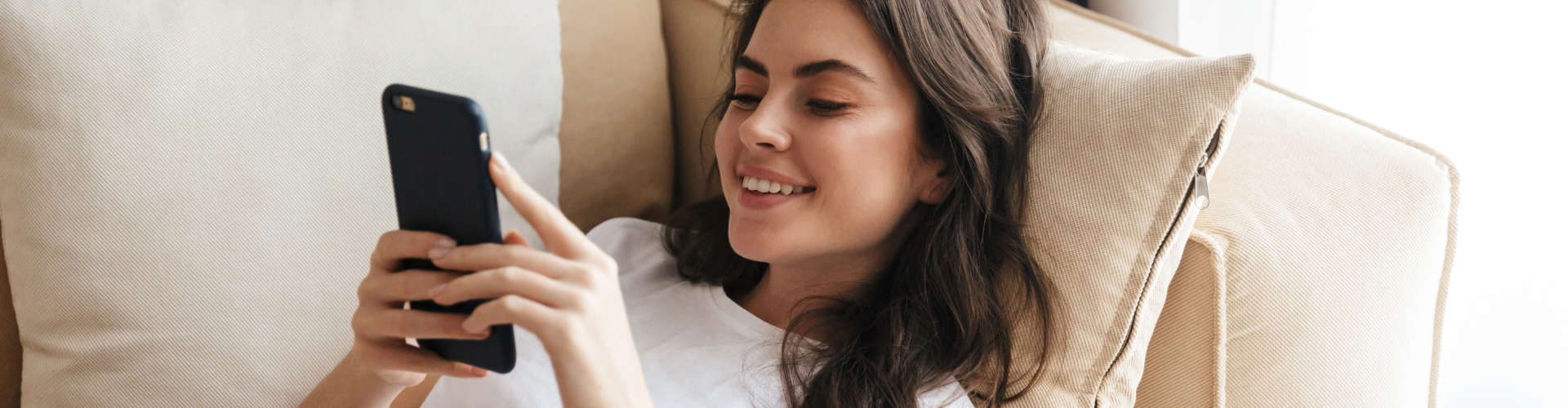 Beautiful smiling young brunette woman relaxing on a couch at home, using mobile phone.
