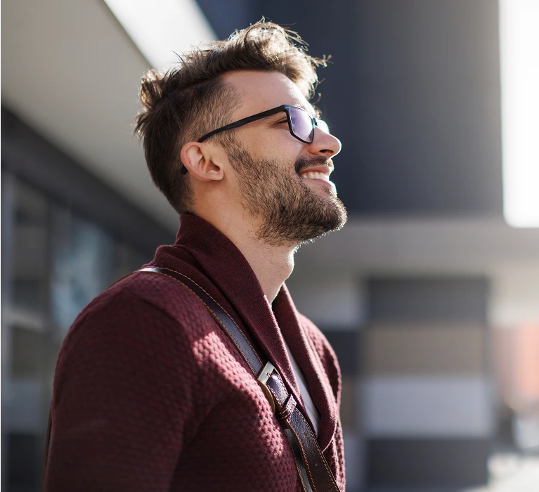 A close-up profile photo of a smiling man with a beard and dark, thick-rimmed glasses, looking up toward the sunlight. He is wearing a burgundy cardigan sweater and has a dark leather strap from a bag across his shoulder. The background is blurred, modern architecture.