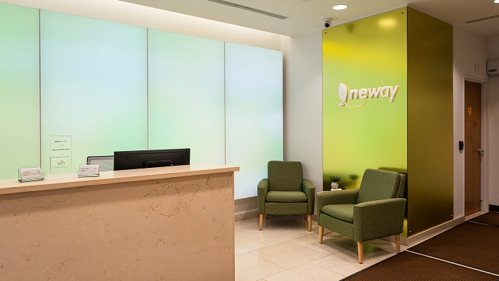 Neways modern reception area with a light beige stone counter on the left and two green upholstered armchairs on the right. Behind the counter is a large, segmented wall display with a gradient of pale green and white light. To the right of the display is a bright, reflective lime-green wall panel featuring the "neway" logo in white and orange.