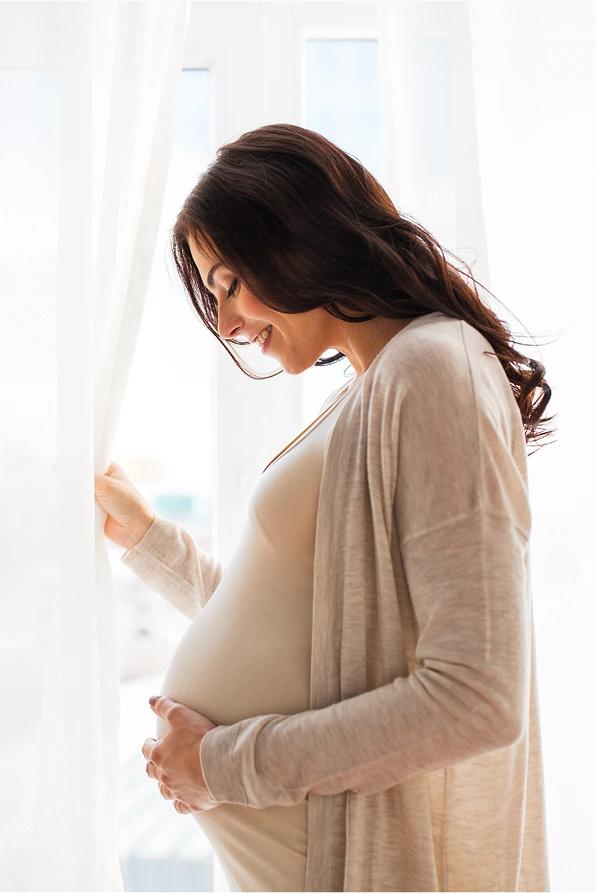A side view of a smiling pregnant woman with long dark hair, standing in front of a bright window. She is holding her bump with both hands and gently pulling aside a white sheer curtain. She is wearing a light-colored dress and a beige cardigan.