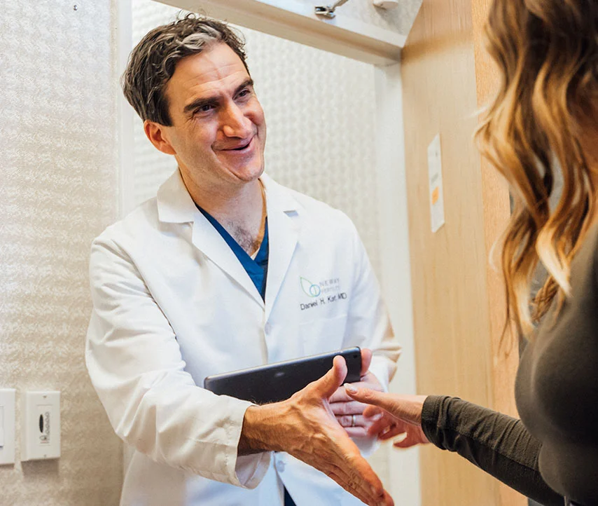 Dr. Kort in a white lab coat, holding a tablet, is shaking hands with a female patient in a doorway. - in New York, NY