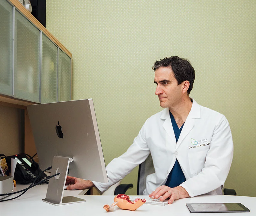 A photo of a doctor, Dr. Kort, sitting at his office desk and working on an Apple desktop computer. He is wearing a white lab coat over blue scrubs. The office has light green walls, wooden cabinets, and a phone on the desk. - in New York, NY
