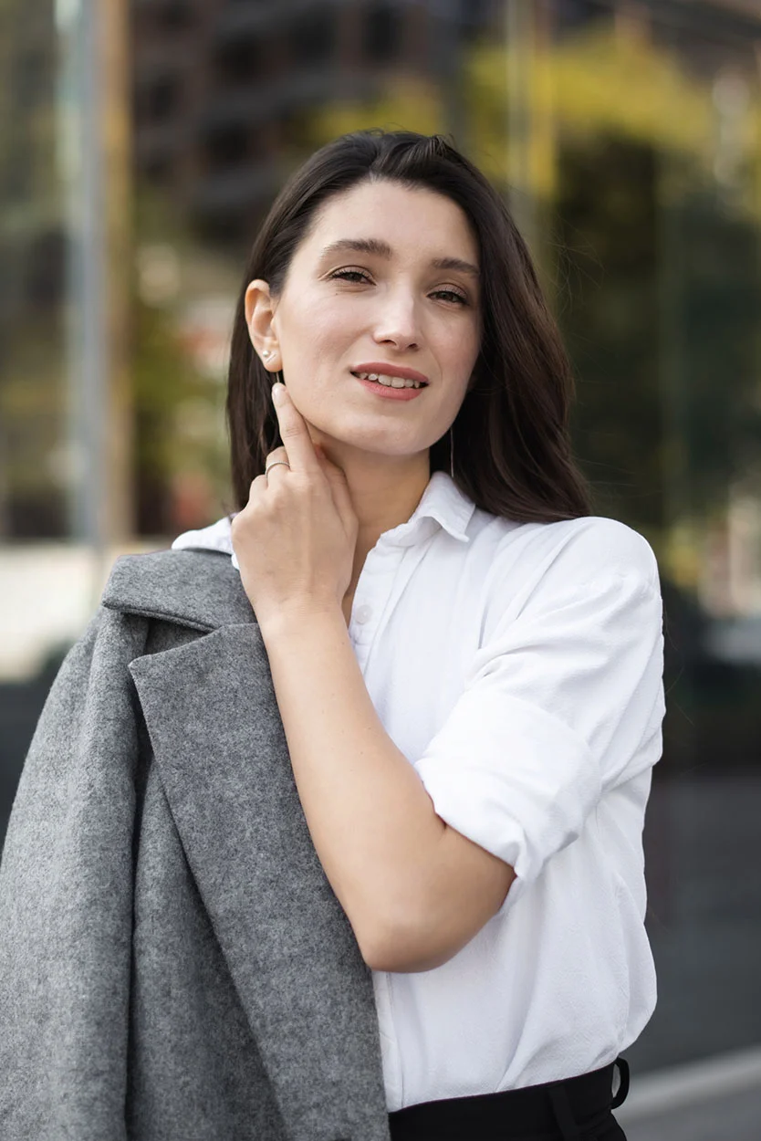 A woman with long dark hair, wearing a white collared shirt, is holding a gray wool coat over her shoulder while gently touching her neck and looking at the camera. - Advanced Maternal Age in New York, NY