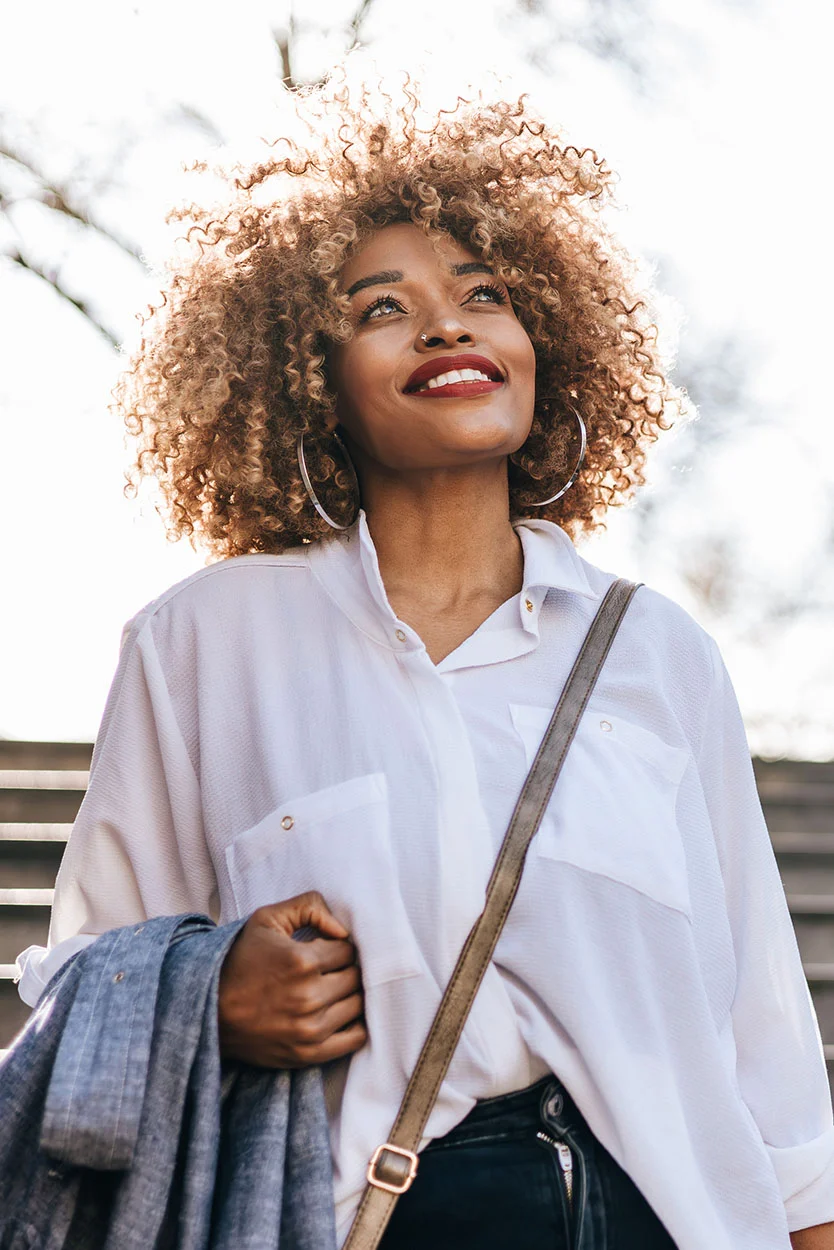 A smiling woman with voluminous curly hair and bright red lipstick is standing outdoors, looking up into the sunlight while wearing a white collared shirt. - Assisted Hatching in New York, NY