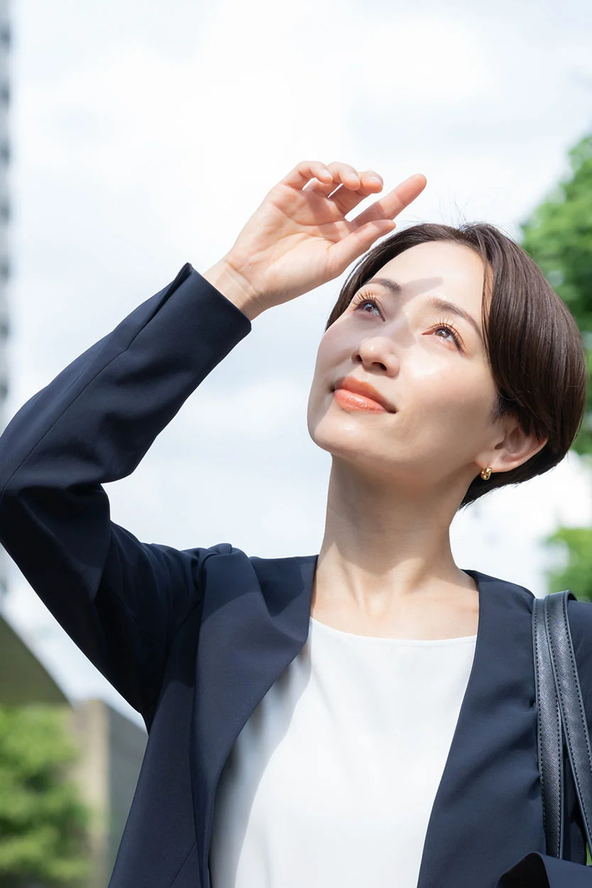 A woman with short dark hair, wearing a navy blazer and white top, is outdoors, shielding her eyes with her hand while looking up at the bright sun. - Blastocyst Culture Transfer in New York, NY