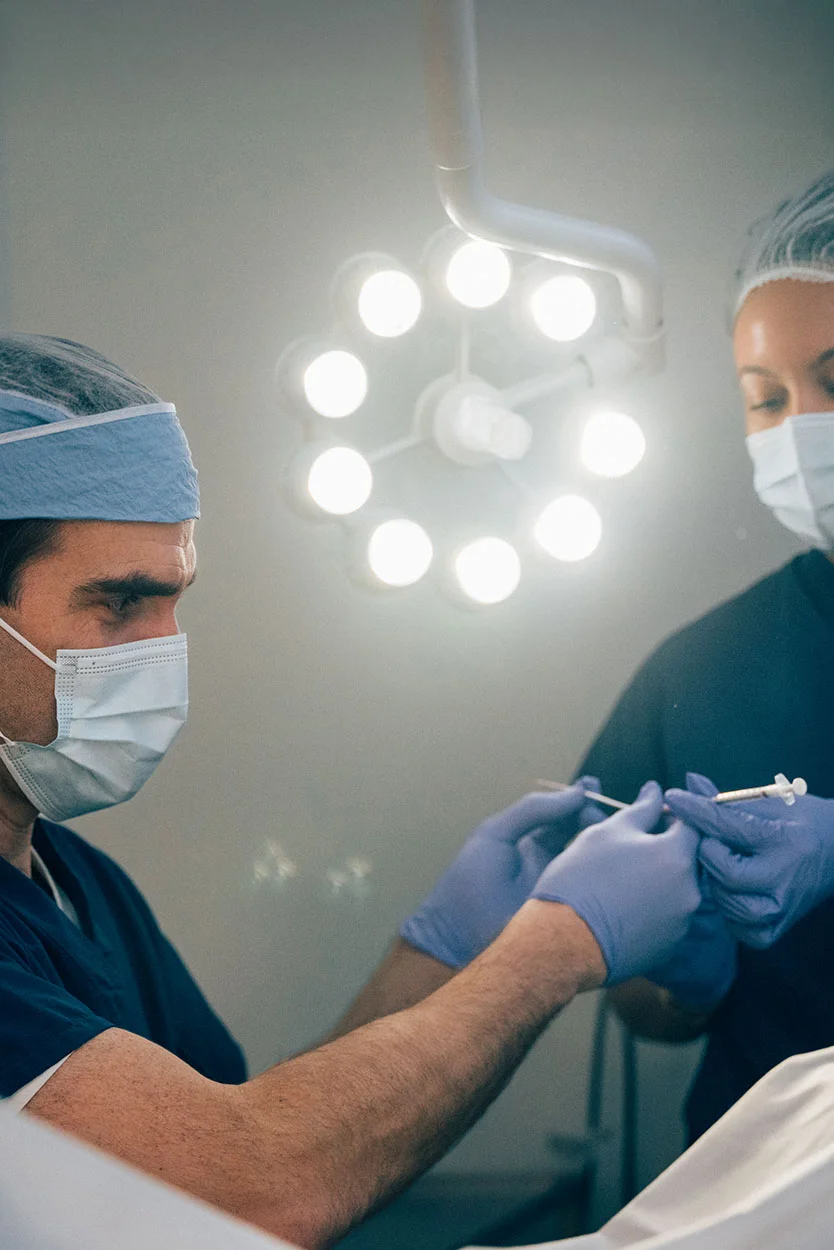 Dr. Kort and surgical assistant in blue scrubs, gloves, and a mask is holding a small medical instrument, possibly a syringe or catheter, while working in an operating room under bright surgical lights. - Elective Single Embryo Transfer in New York, NY