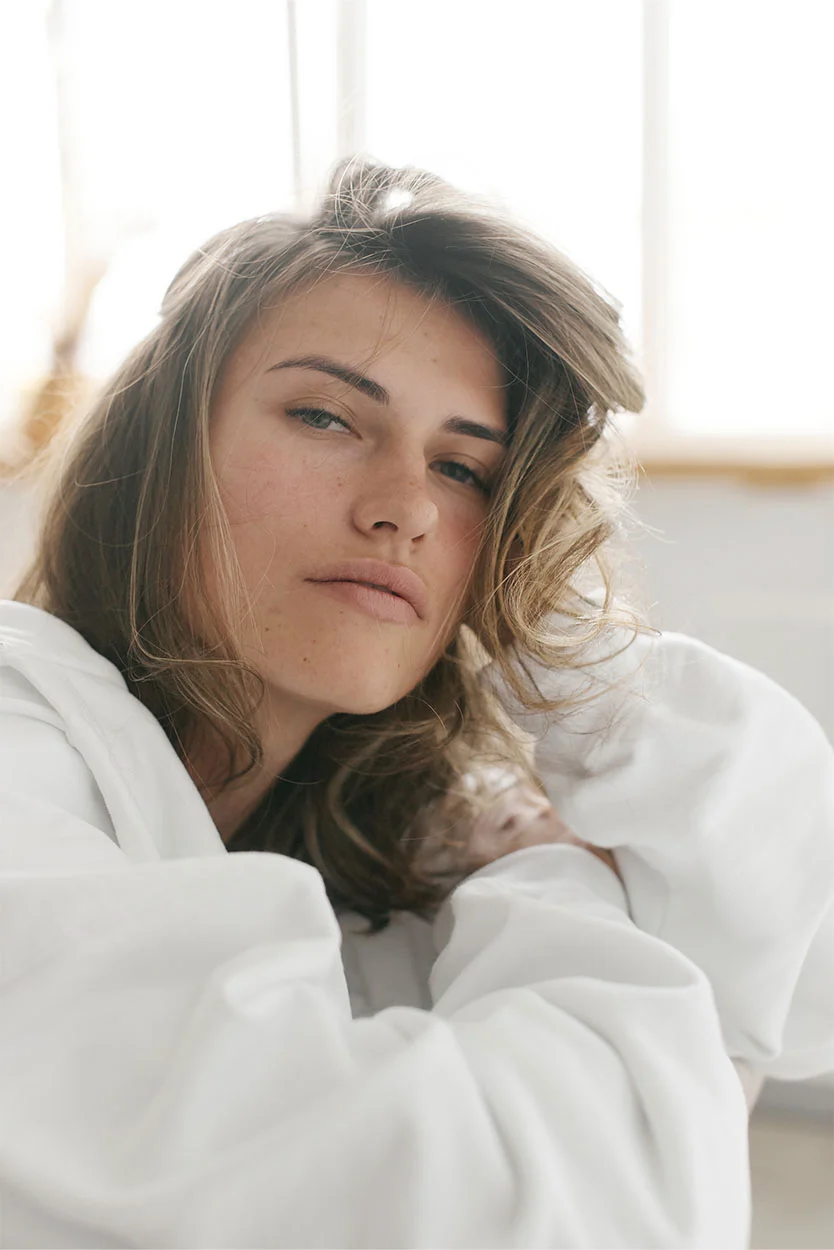 A woman with shoulder-length brown hair, wearing a white oversized shirt, is looking intently at the camera in a brightly lit room. - Endrometriosis in New York, NY