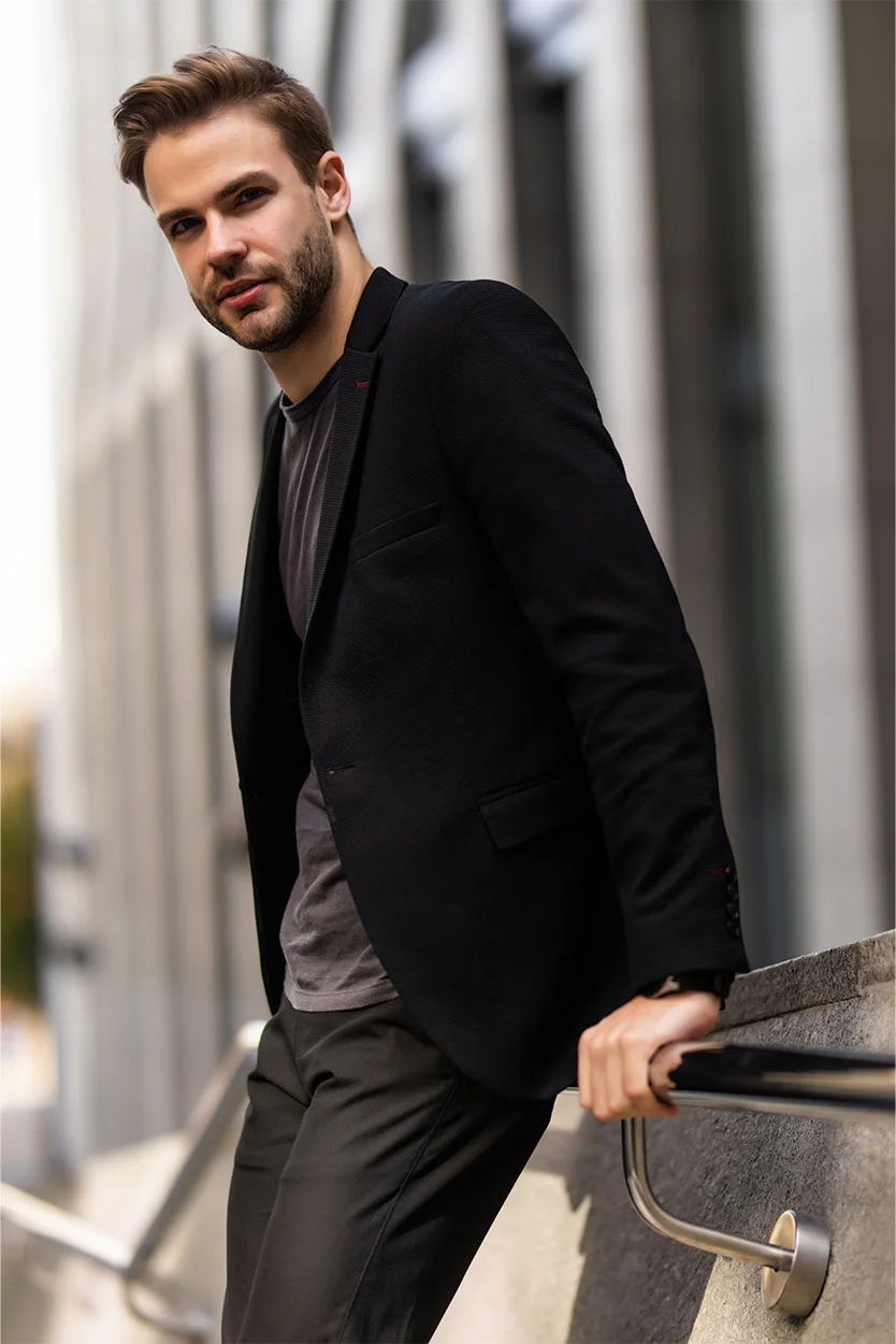 A stylish man with a beard and light brown hair, wearing a black blazer over a gray t-shirt, is leaning on a railing outdoors, looking at the camera. - Enlarged Scrotu Veins Varicocele in New York, NY