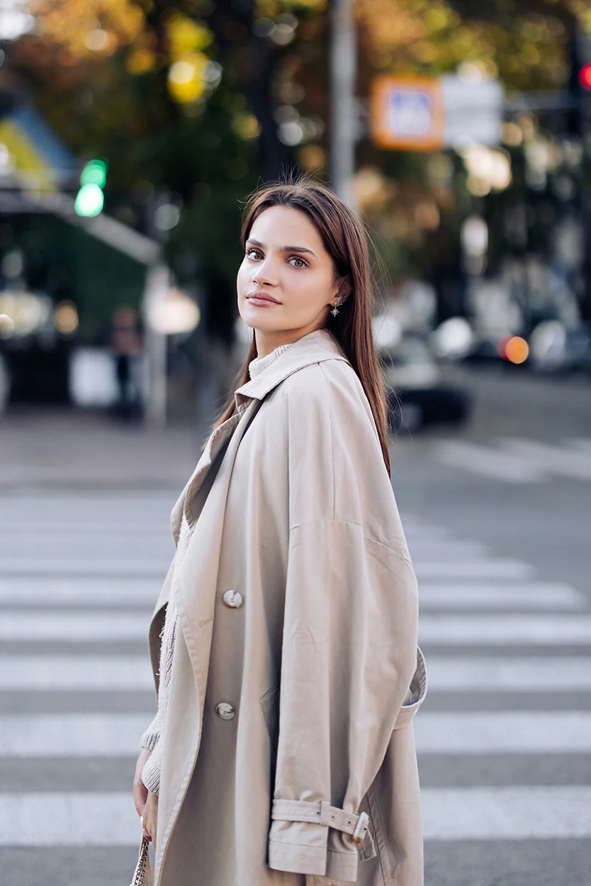 A woman with long dark hair, wearing a loose beige trench coat and a light turtleneck, is standing in a city crosswalk and looking over her shoulder. - Fallopian Tube Damage or Blockages in New York, NY