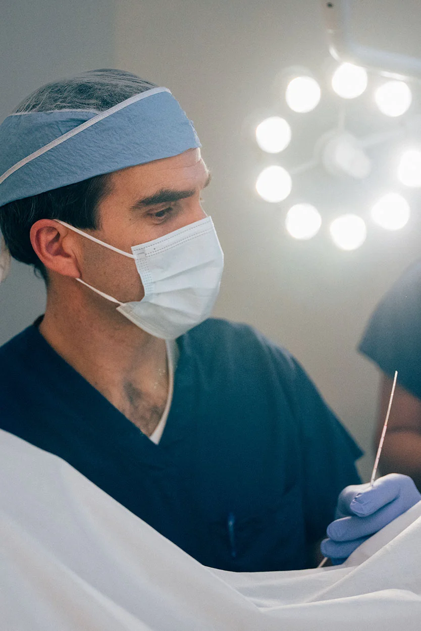 Dr. Kort in a blue surgical cap, mask, and scrubs is holding a thin medical instrument while working under bright surgical lights. - Gestational Surrogacy in New York, NY