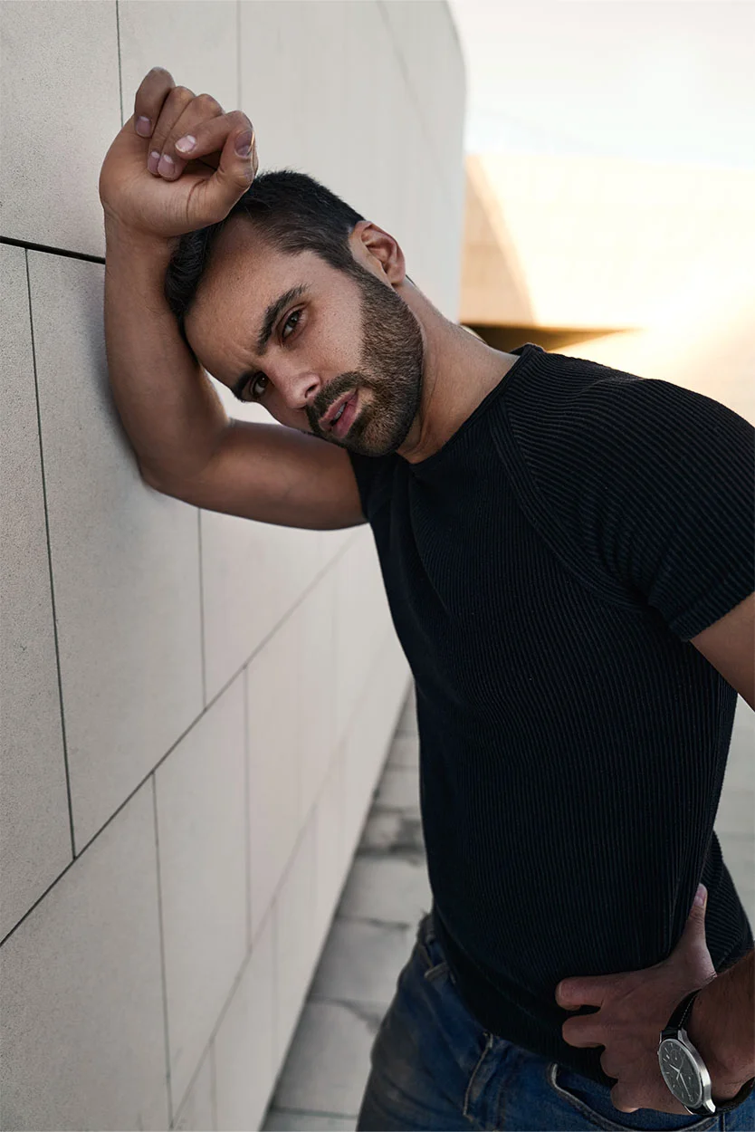 A man with dark hair and a beard, wearing a black textured t-shirt and jeans, is leaning against a light-colored wall outdoors, looking directly at the camera. - High Heat Exposure to Testicles in New York, NY