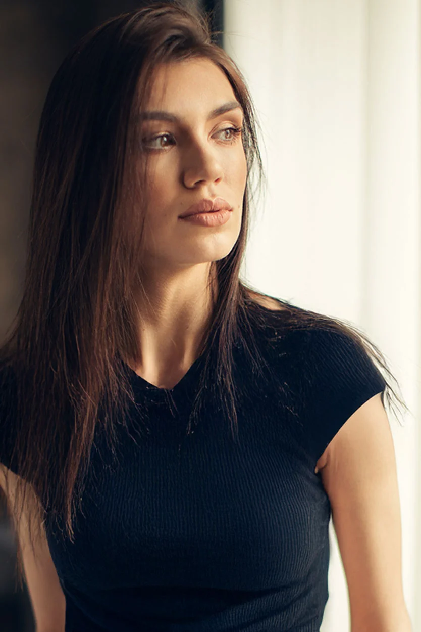 A woman with long dark hair, wearing a black top, is looking thoughtfully out a window with soft light on her face. - Hyperpitutarism in New York, NY