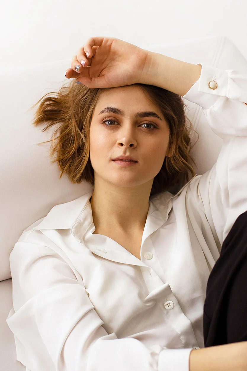 A portrait of a woman with short brown hair, wearing a white collared shirt and black pants, lying on a white surface with one arm resting on her forehead. - Hyperprolactinemia in New York, NY