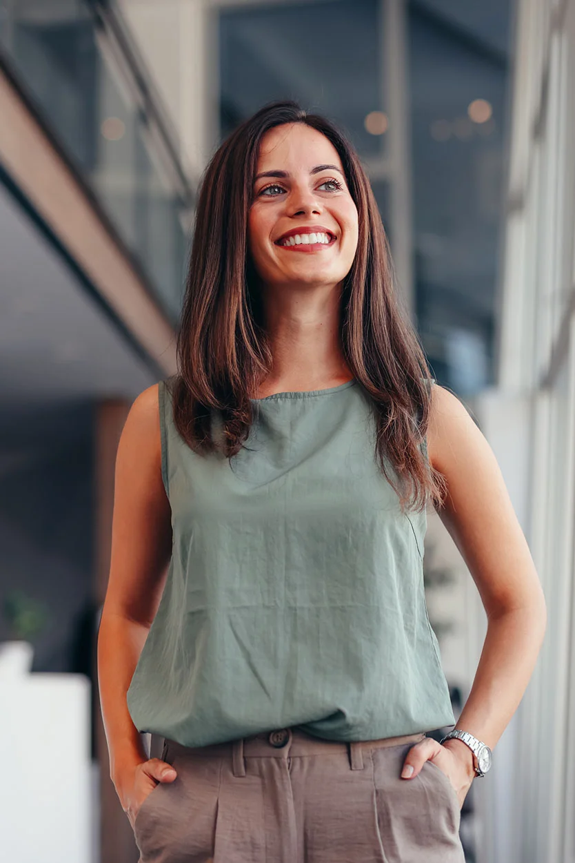 A smiling young woman with brown hair and a green sleeveless top stands indoors with her hands in the pockets of her brown pants, looking up and to the side. - Implantation Failure in New York, NY