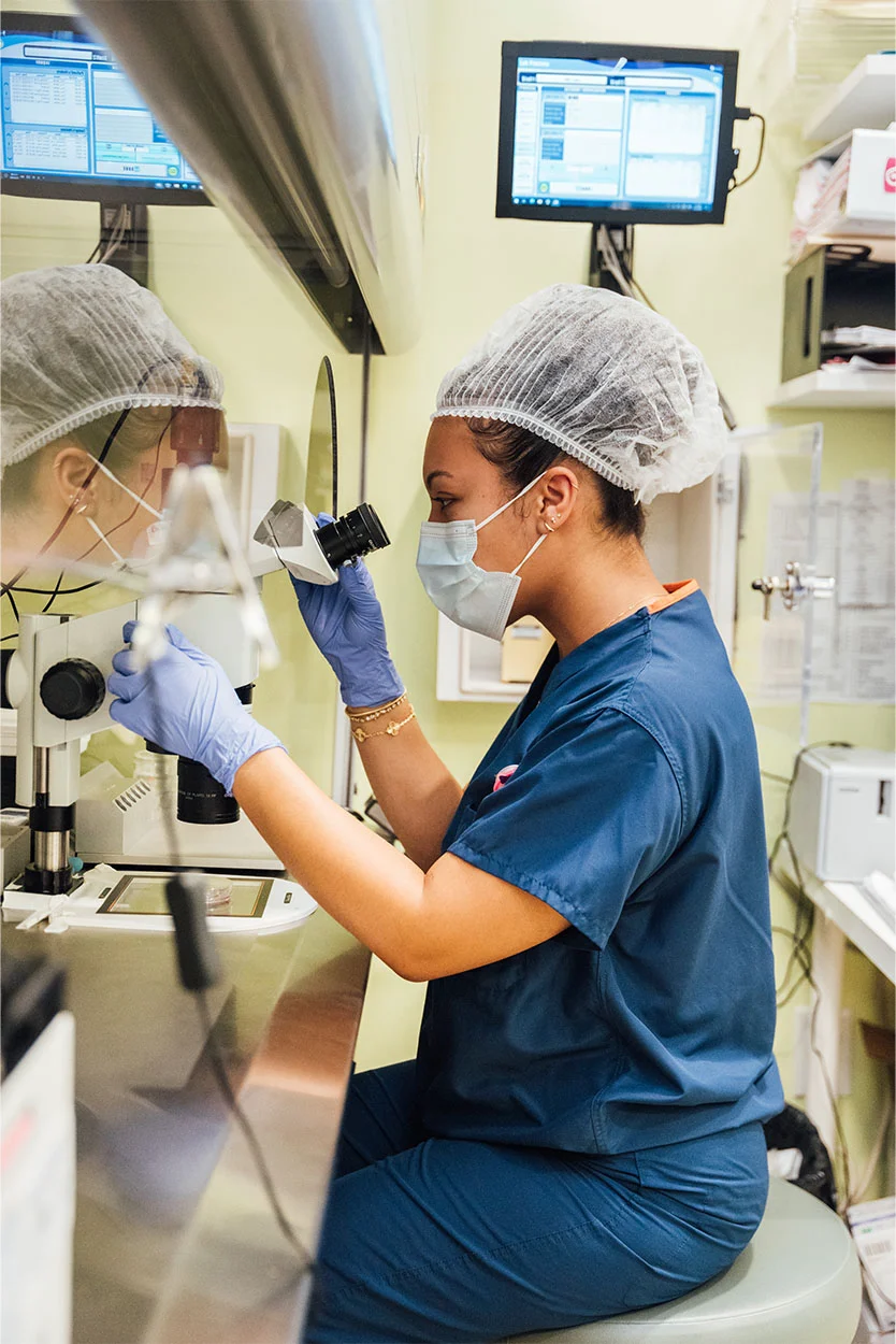 A female lab technician wearing a hair net and face mask is looking through a microscope in a brightly lit laboratory. She is wearing blue scrubs, and computer screens and white shelves are visible in the background against a light green wall. - In Vitro Maturation IVM in New York, NY