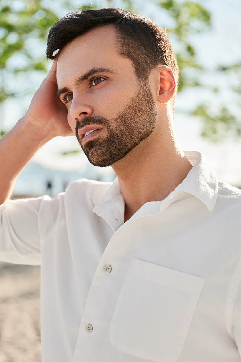 A man with a beard and dark hair wearing a white collared shirt stands outside, looking off to the side with one hand resting near his head. - Injury to the Scrotum or Testicles in New York, NY