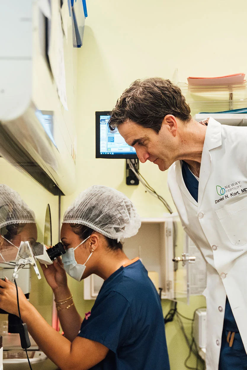 Dr. Kort, wearing a white lab coat, bends over to look at a female technician who is working at a microscope inside a laboratory hood. The bright lab setting has light green walls and shelves with files and schedules. - Intractoplasmatic Sperm Injection ICSI in New York, NY