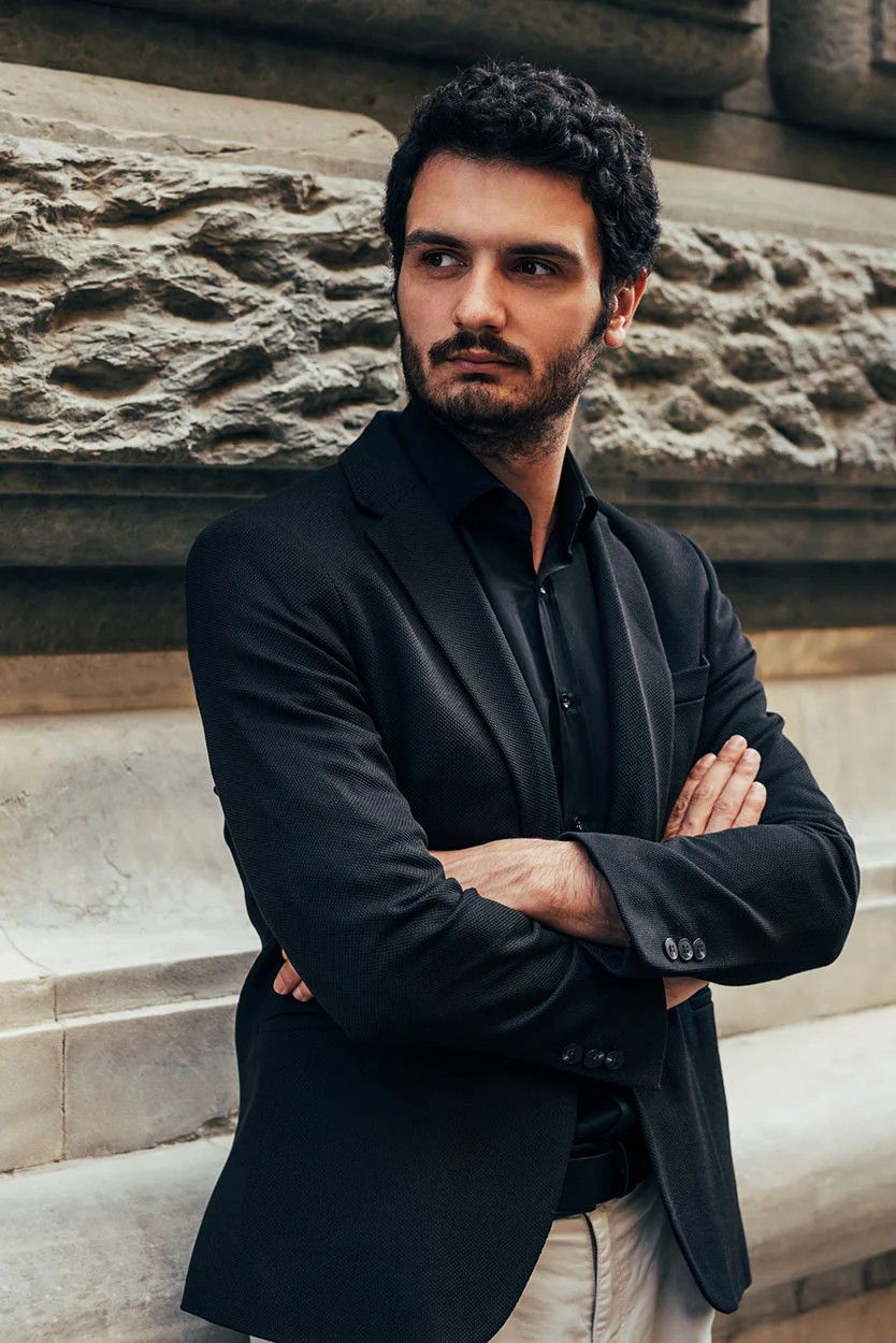 A man with dark, curly hair and a beard, wearing a black blazer and black shirt, stands with his arms crossed outside against a stone wall. He is looking off to the side with a serious expression. - Low Sperm Count or Low Testosterone Hypogonadism in New York, NY