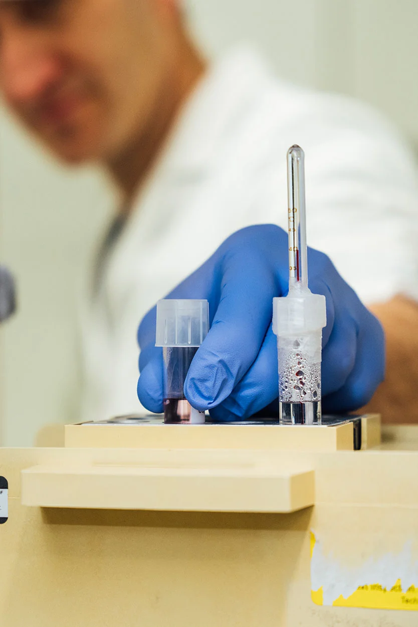 A close-up shot of a gloved hand holding a small test tube containing a dark liquid in a laboratory setting. Another small vial with a thermometer or capillary tube is visible next to it. The person wearing the lab coat is blurred in the background. - Mild IVF in New York, NY