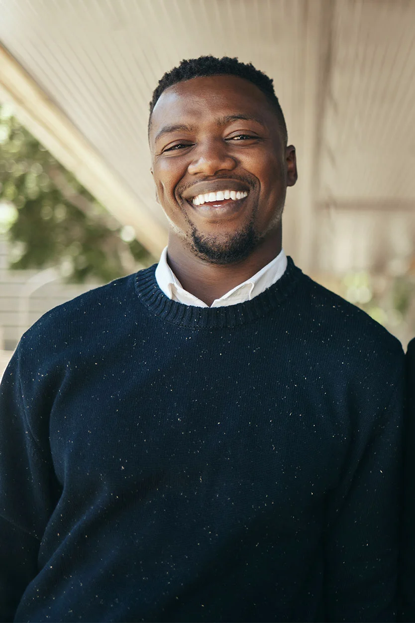 A bright vertical portrait of a smiling man with short dark hair and a beard. He is wearing a dark blue or black speckled sweater over a white collared shirt. He is standing outdoors under a covered porch. - Misuse or Anabolic Steroids in New York, NY