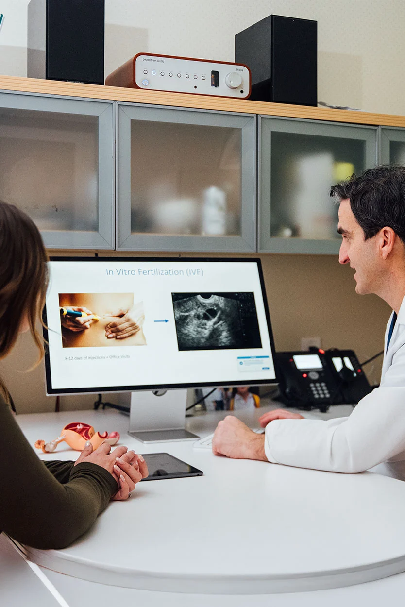 A doctor, Dr. Kort, wearing a white coat, is sitting at a desk and gesturing towards a computer monitor for a patient. The monitor displays images of embryo development labeled "Fertilization (IVF)." The patient is seen in the foreground from behind. - Natural Cycle IVF in New York, NY
