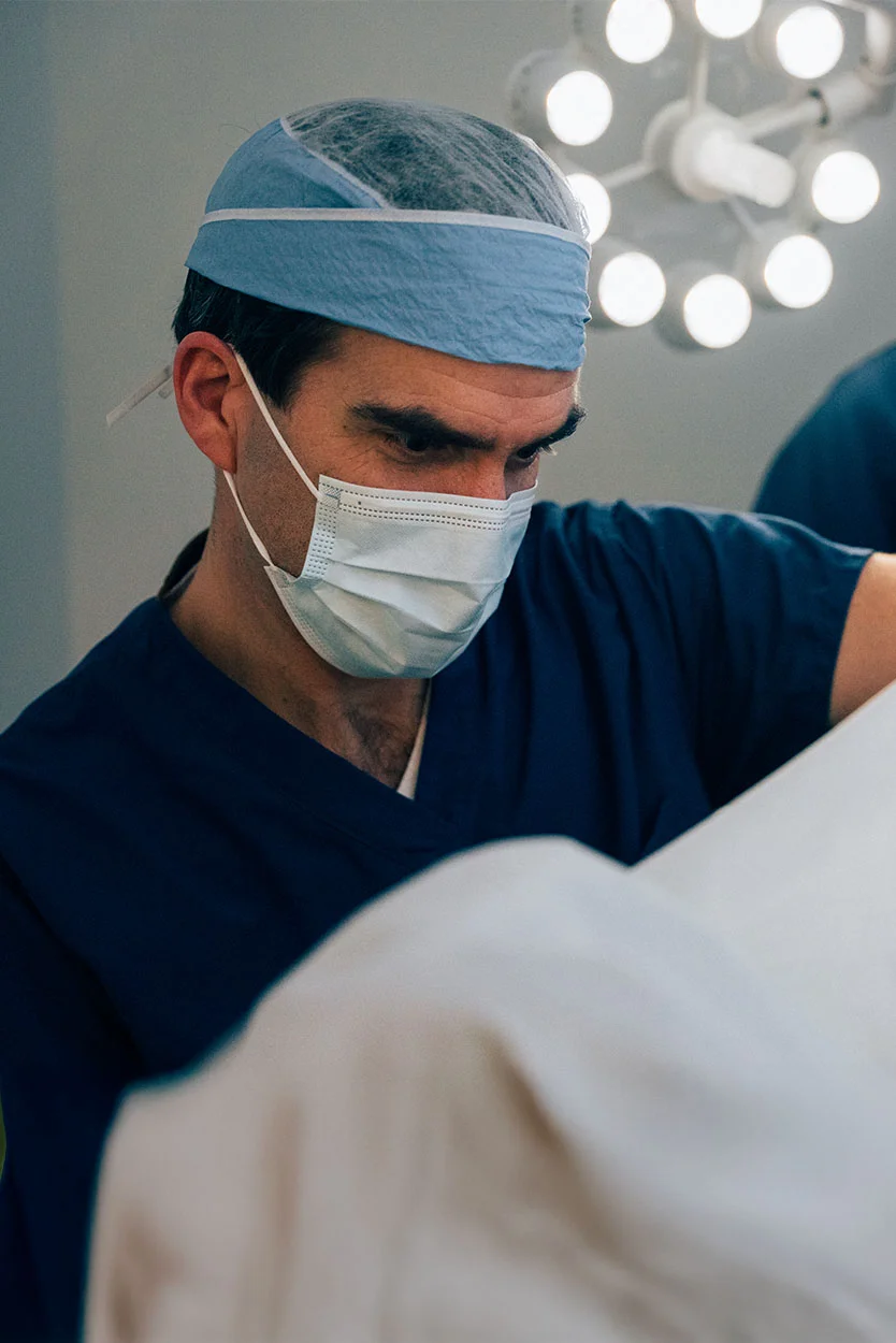 A surgeon wearing blue scrubs, a surgical cap, and a face mask is looking down intently, with a bright operating room light fixture visible in the blurred background. - Polyps in the Uterus in New York, NY