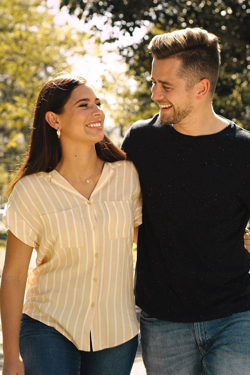 A smiling couple walking outdoors in a sunny park setting. The woman on the left is wearing a beige and white striped shirt and blue jeans, looking up at the man on the right, who is wearing a black shirt and has his arm around her. - Preimplantation Genetic Screening PGS in New York, NY