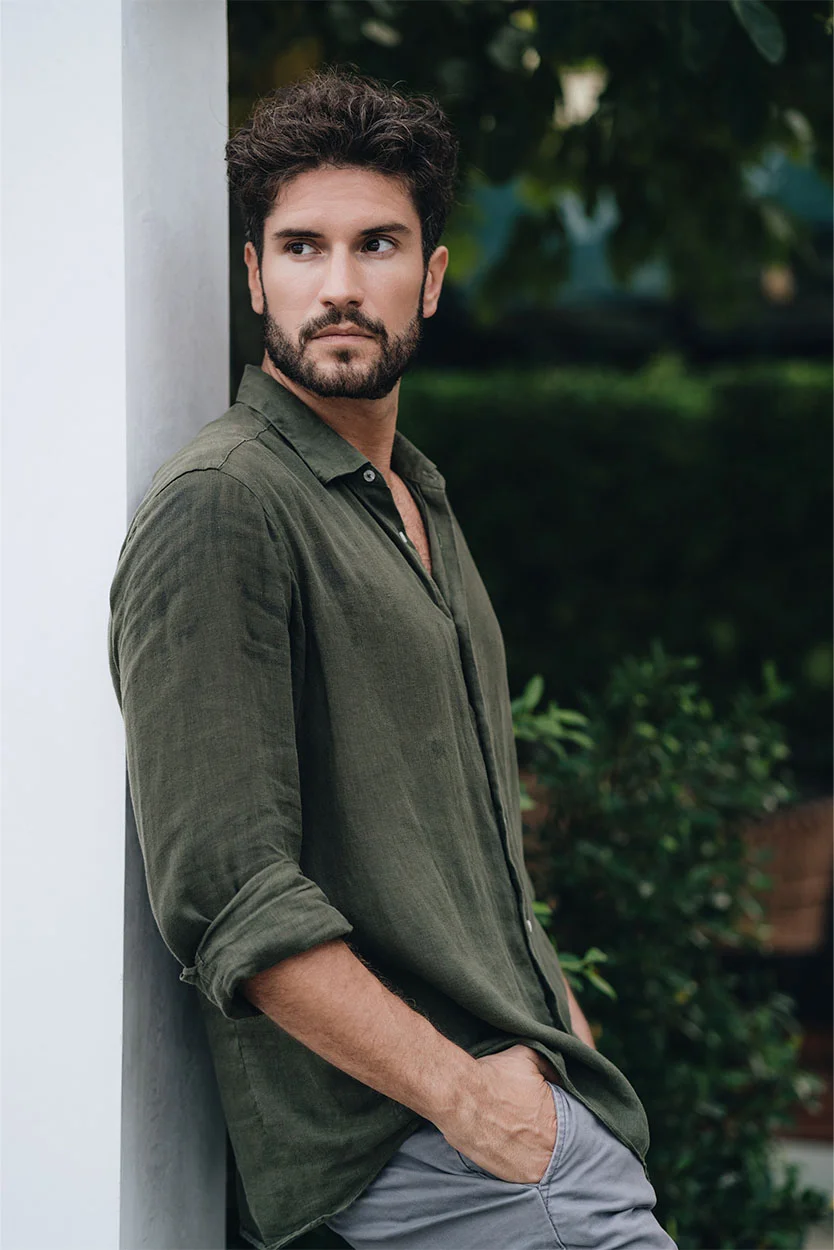 A portrait of a man with dark, curly hair and a beard, leaning against a white pillar outdoors. He is wearing an olive-green linen shirt with the sleeves rolled up and gray trousers, looking off to the side. - Premature Ejaculation or Retrograde Ejaculation in New York, NY