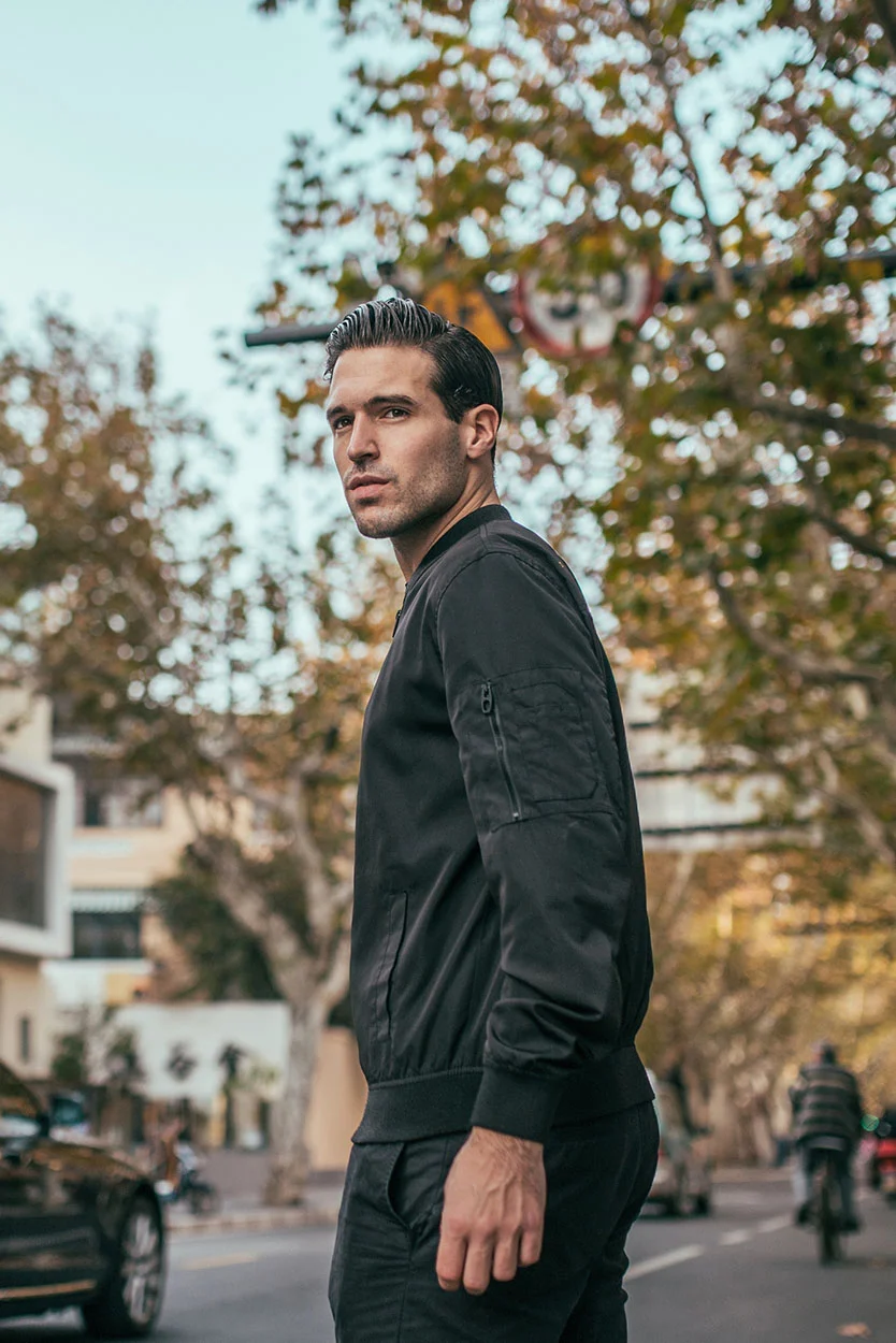 A handsome man with dark hair and a black bomber jacket is standing on a city street, looking over his shoulder. - Semen Analysis in New York, NY