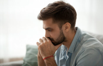 Man sitting indoors, appearing deep in thought, with hands clasped near his face. Wearing a denim shirt and a red string bracelet.