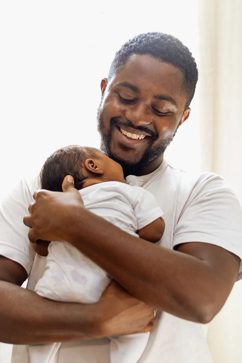 A happy Black man with a beard and short dark hair, wearing a white t-shirt, is gently holding a newborn baby dressed in white, who is resting its head against his shoulder. He is smiling with his eyes closed, showing affection for the baby, against a bright, light background. - Single dad to be in New York, NY