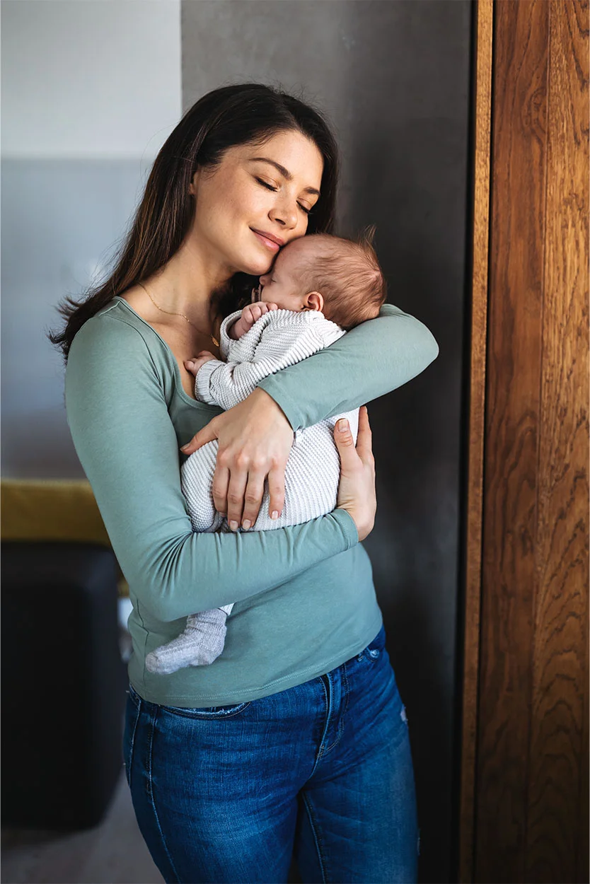 A serene woman with long dark hair, wearing a light green long-sleeve shirt and jeans, is gently holding a peacefully sleeping infant against her chest, with her eyes closed. - Single mom to be in New York, NY