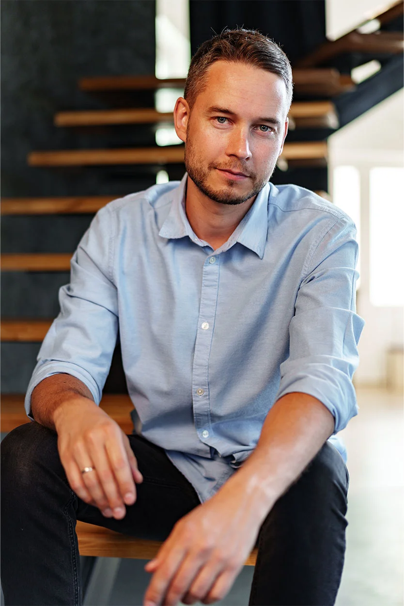 A man with short brown hair and a light beard is sitting on wooden stairs, wearing a light blue collared shirt and black pants, looking directly at the camera. - Testicular Cancer in New York, NY