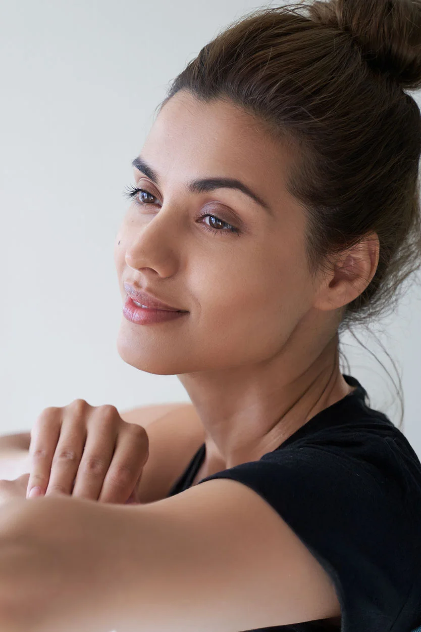 A smiling woman with her hair in a bun is looking to the side, wearing a black shirt, with her face softly illuminated against a light background. - Thyroid Hormone Imbalances in New York, NY