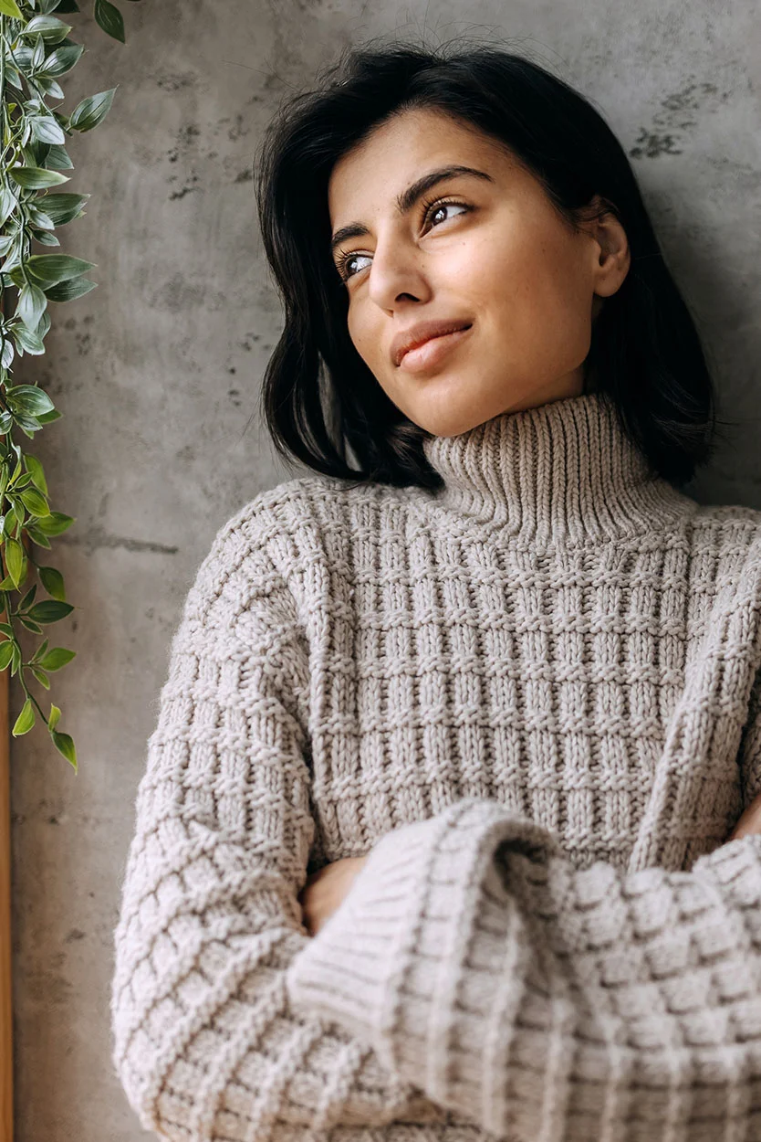 A young woman with dark hair, wearing a cozy, patterned beige turtleneck sweater, is leaning against a concrete wall and looking thoughtfully to the side. - Turners Syndrome in New York, NY