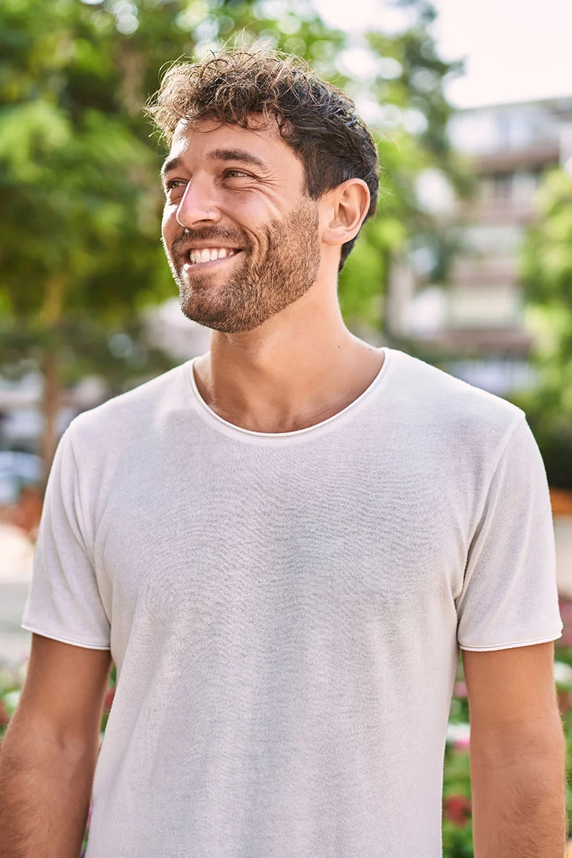 A smiling man with curly brown hair and a beard is wearing a white t-shirt, standing outdoors with trees in the background. - Undescended Testicles in New York, NY