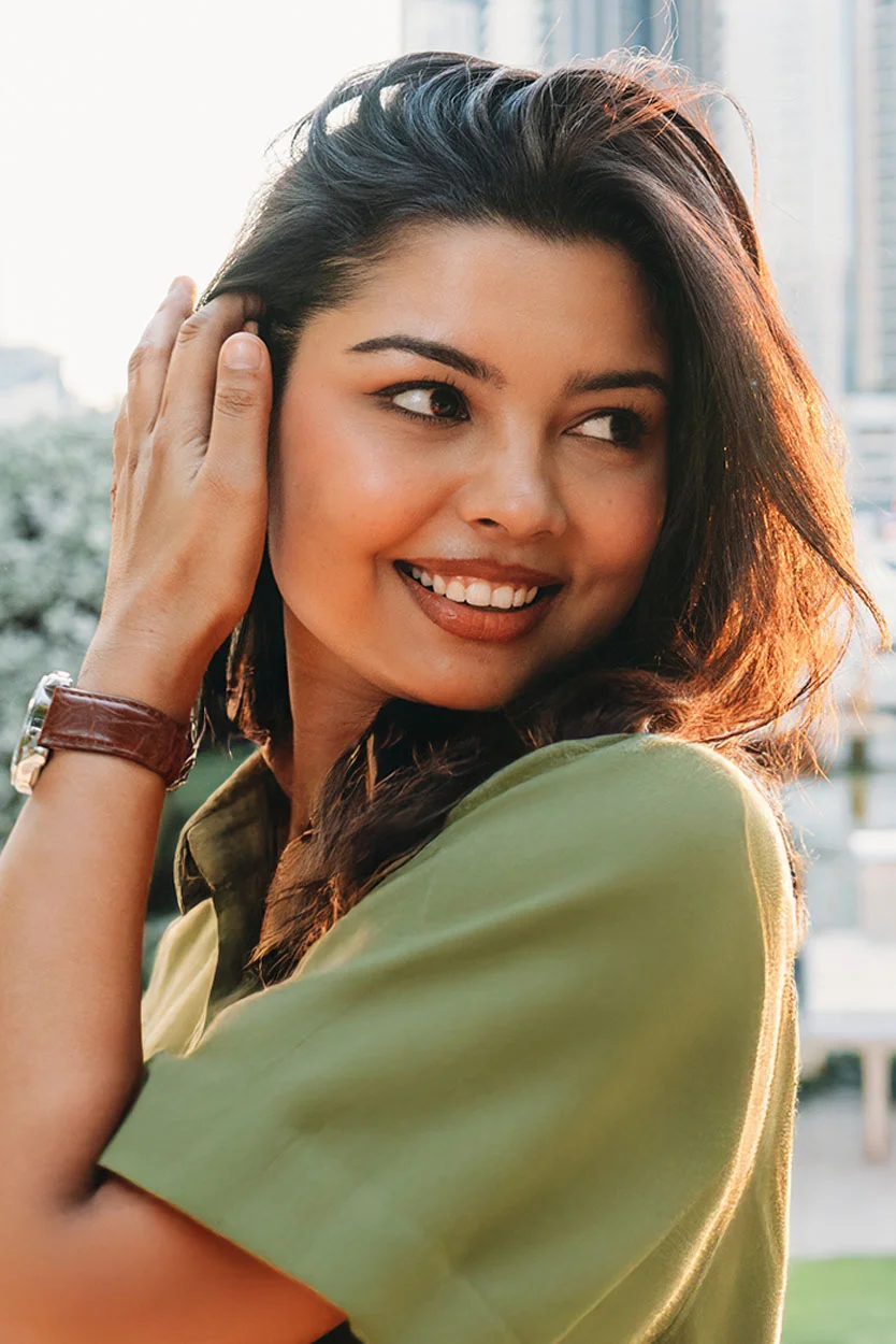 A smiling woman with dark hair and a brown wrist watch, wearing a green shirt, is looking away from the camera in a sunlit outdoor setting. - Uterine or Cervical Abnormalities in New York, NY